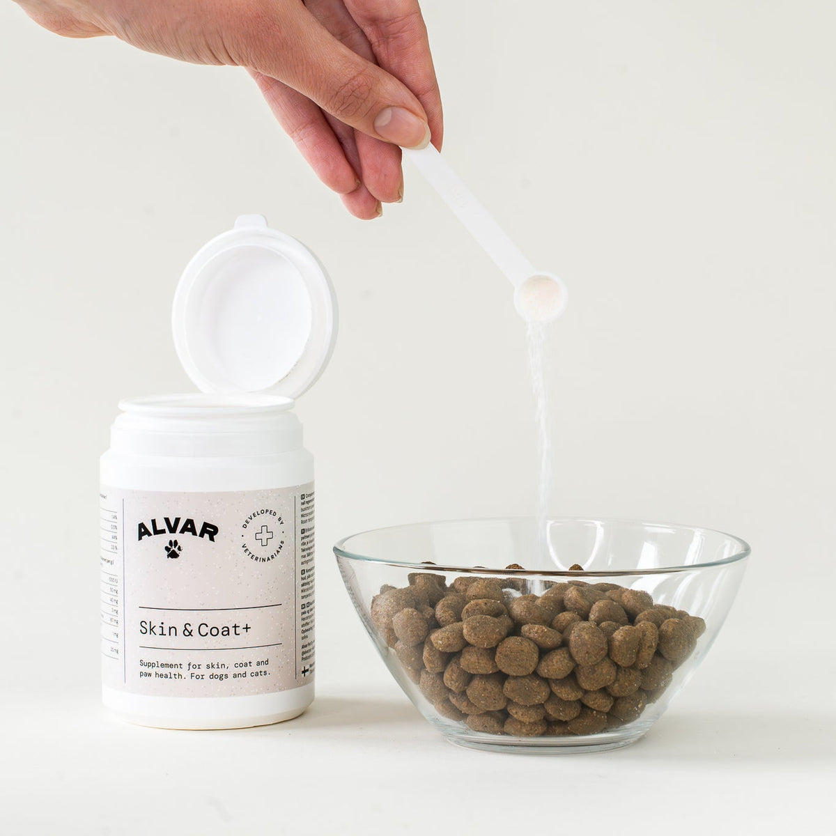 Person holding a spoon over a bowl of cat food next to an open Alvar Skin & Coat supplement bottle.