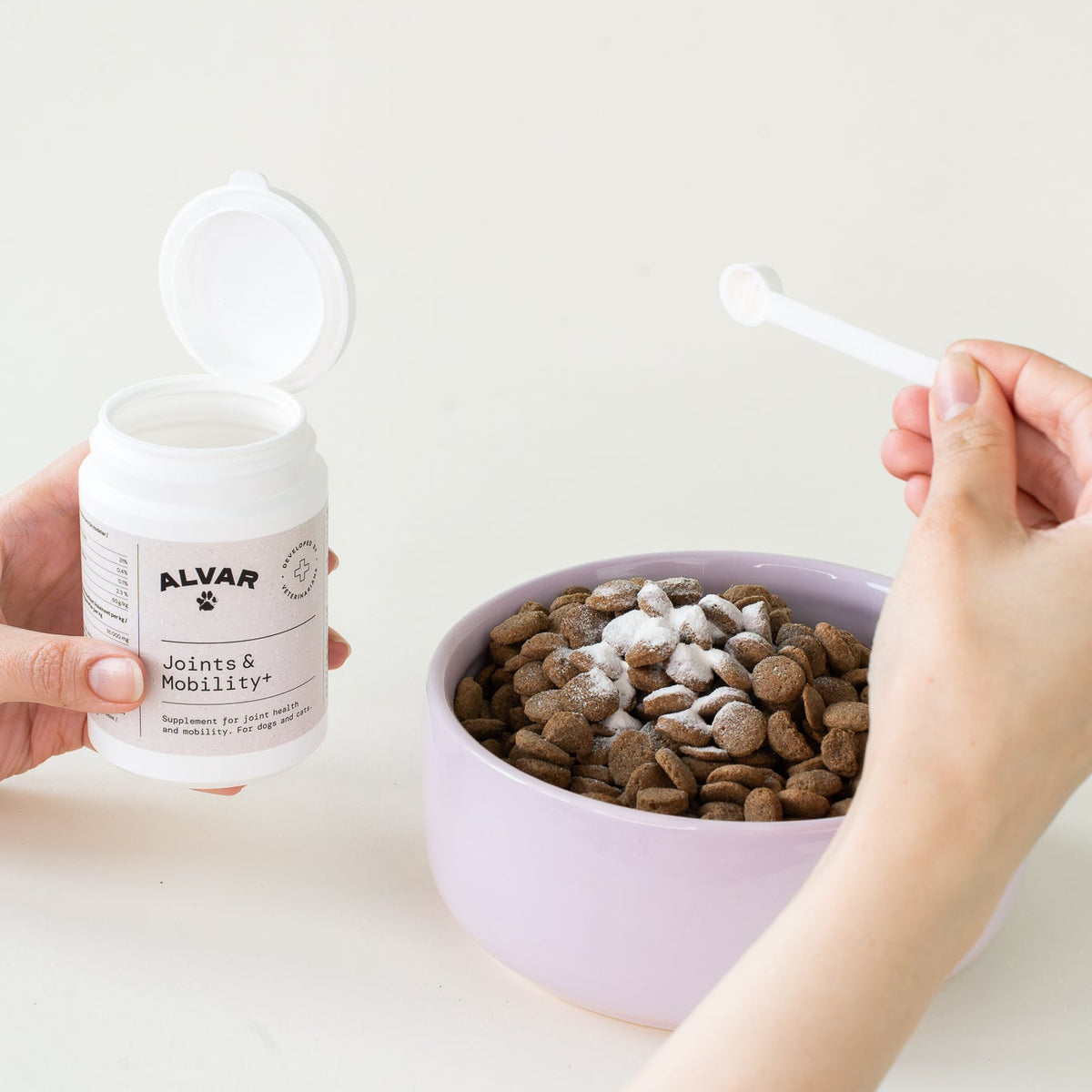Person holding a container of Alvar pet supplement next to a bowl of pet food on a white surface.